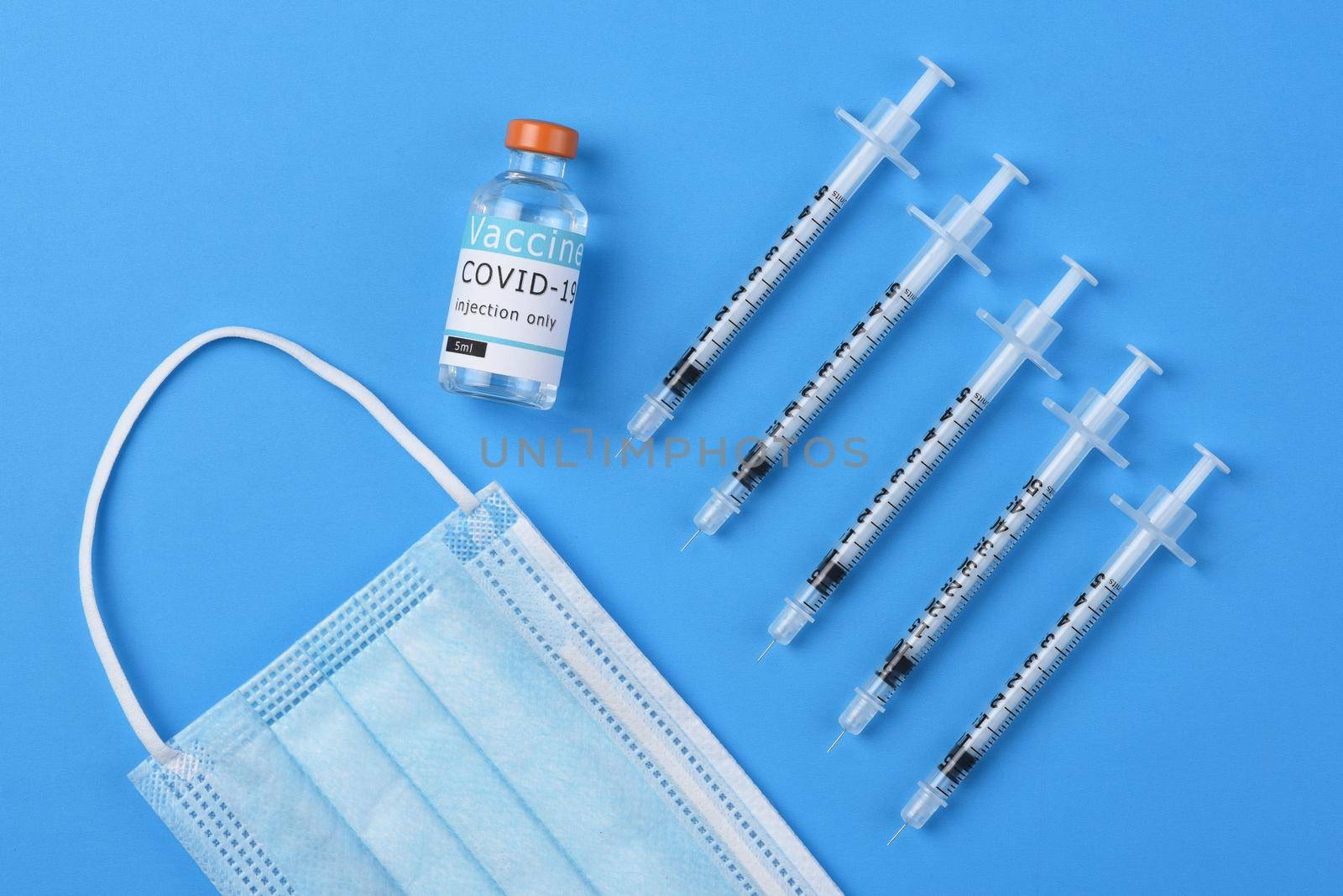 Flat lay shot of syringes a vial of Covid-19 vaccine and a surgical mask on a blue background.