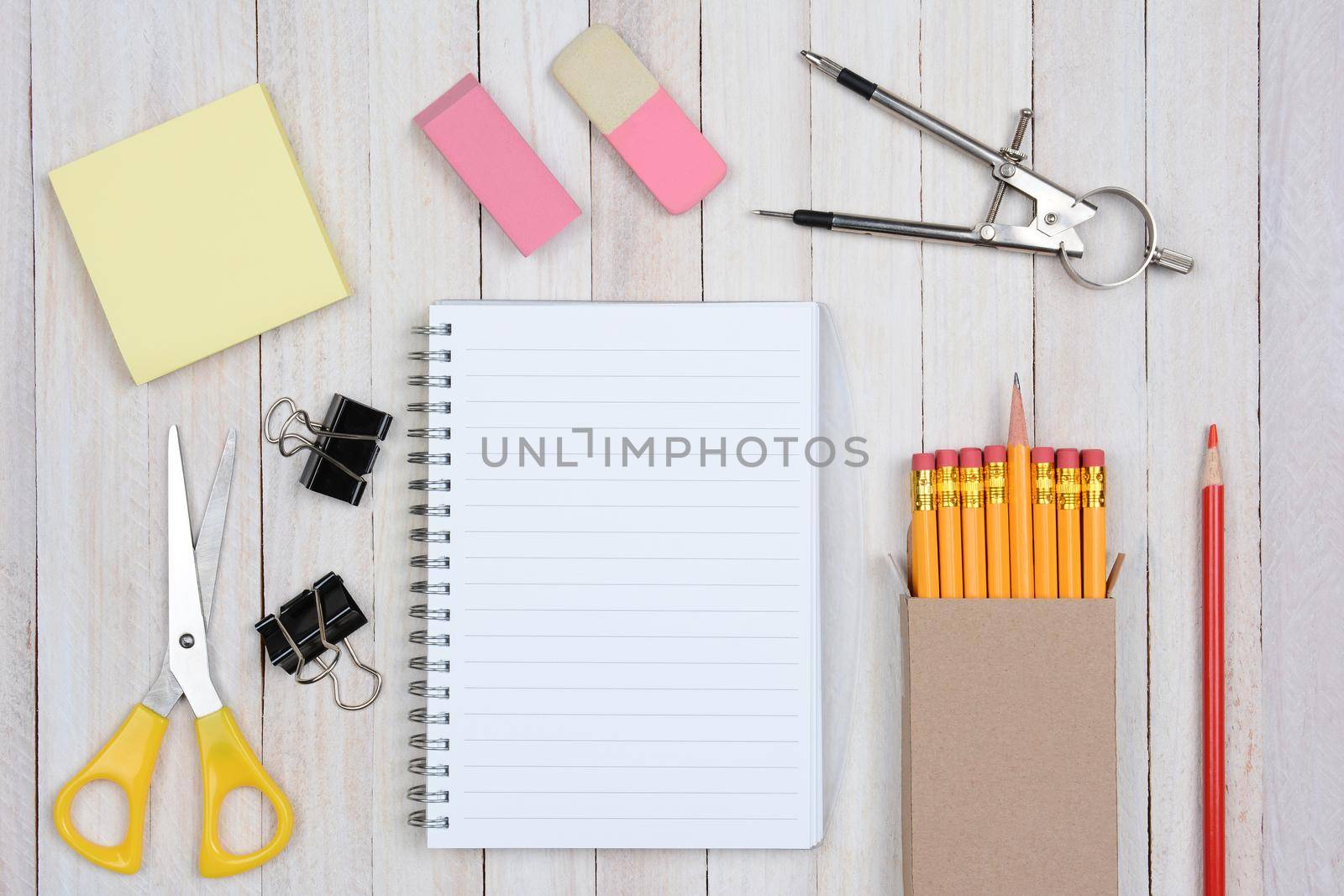A group of items typically found in a school desk. Items include: erasers, pencils, compass, scissors, paper, note pad, paper clips, shot from a high angle.