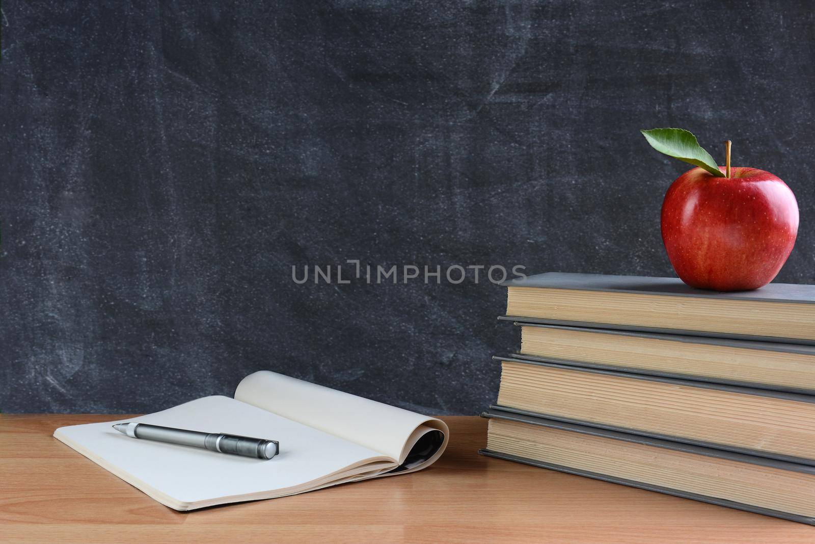 Closeup of a teachers desk with books, paper and pen and a red apple in front of a chalkboard. Horizontal format with copy space. 