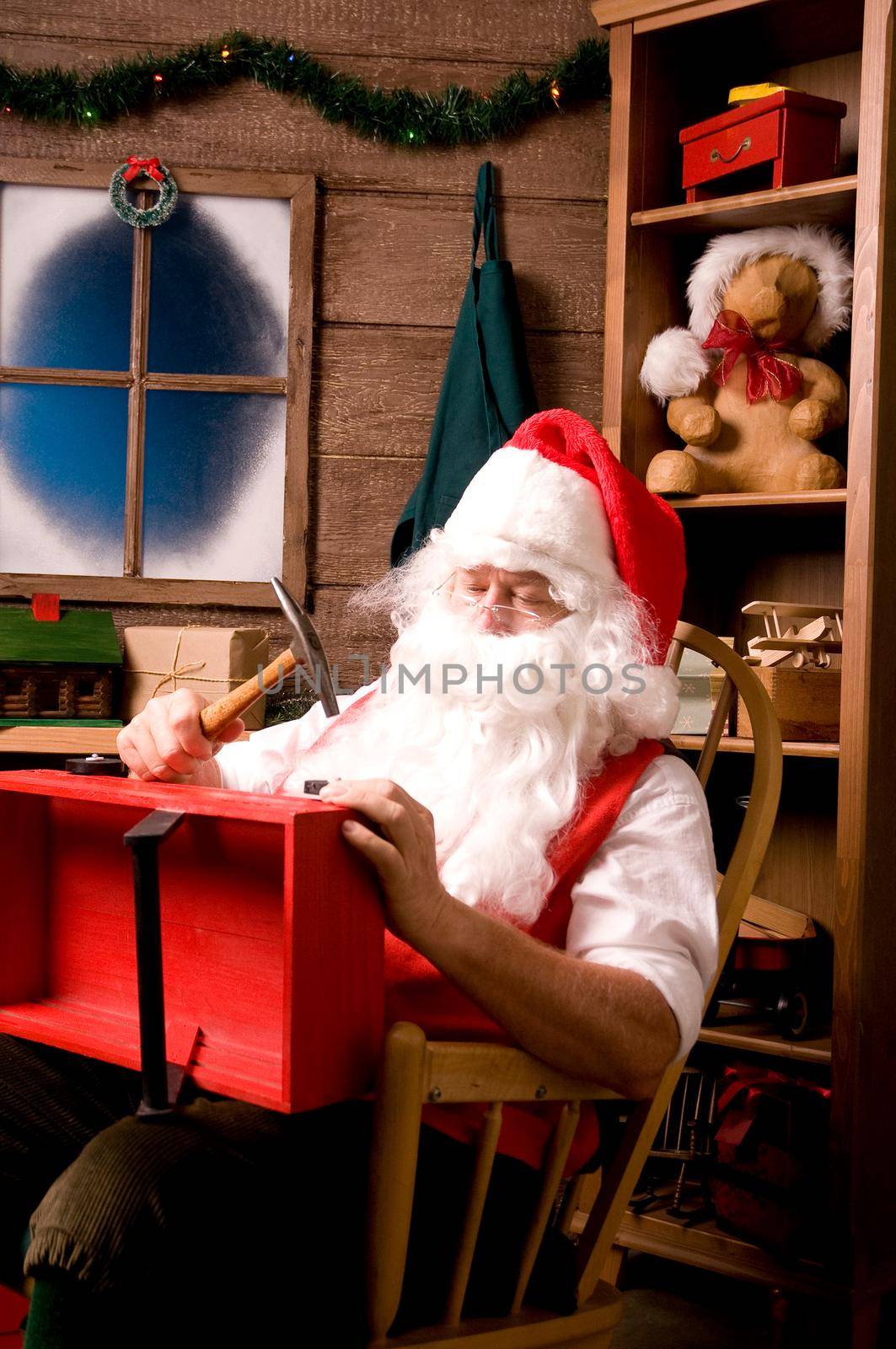 Santa Claus sitting in Rocking Chair in Workshop With Red wooden Wagon on his Lap, Vertical Composition