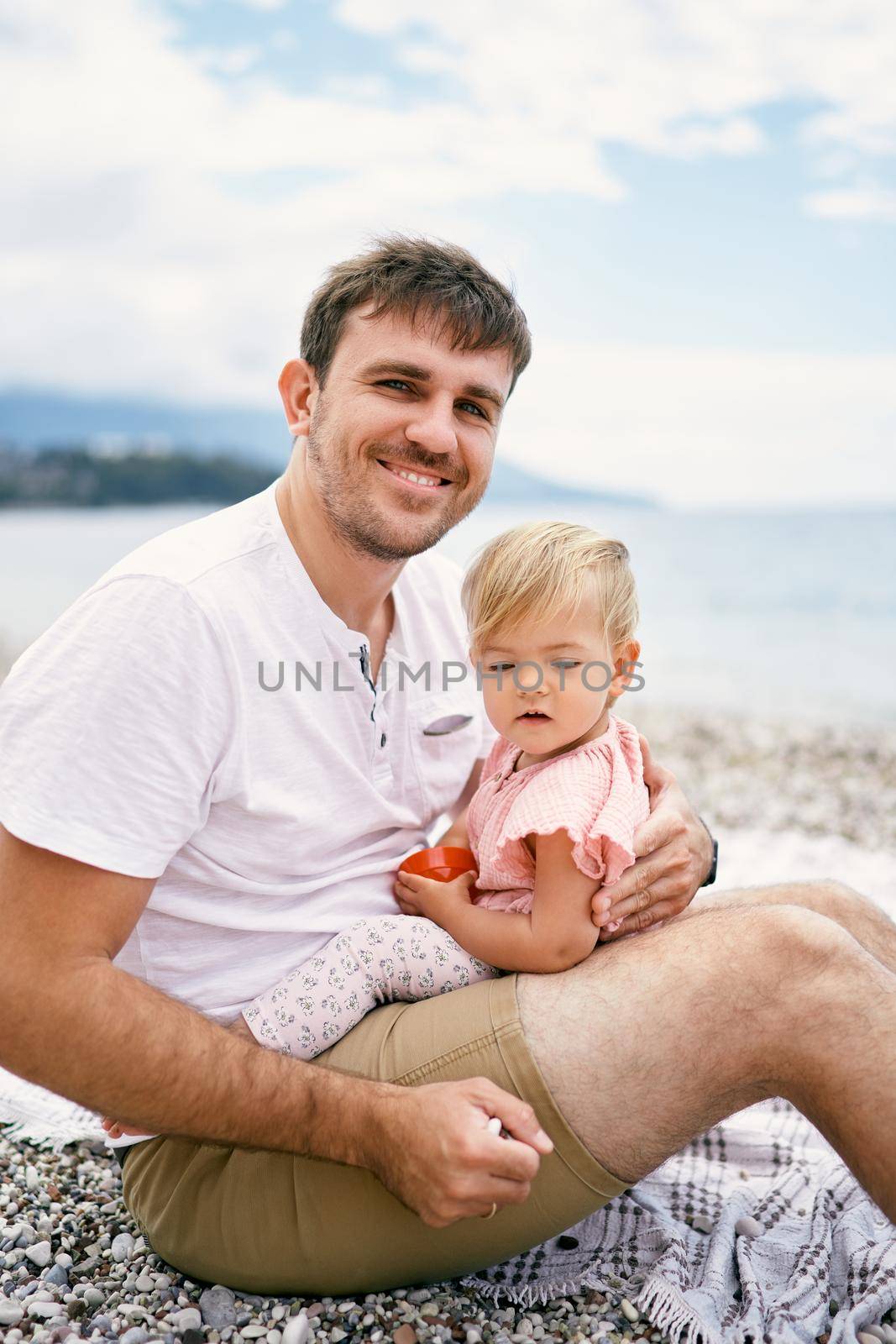 Smiling dad holding a little girl in his arms sitting on a pebble beach by Nadtochiy