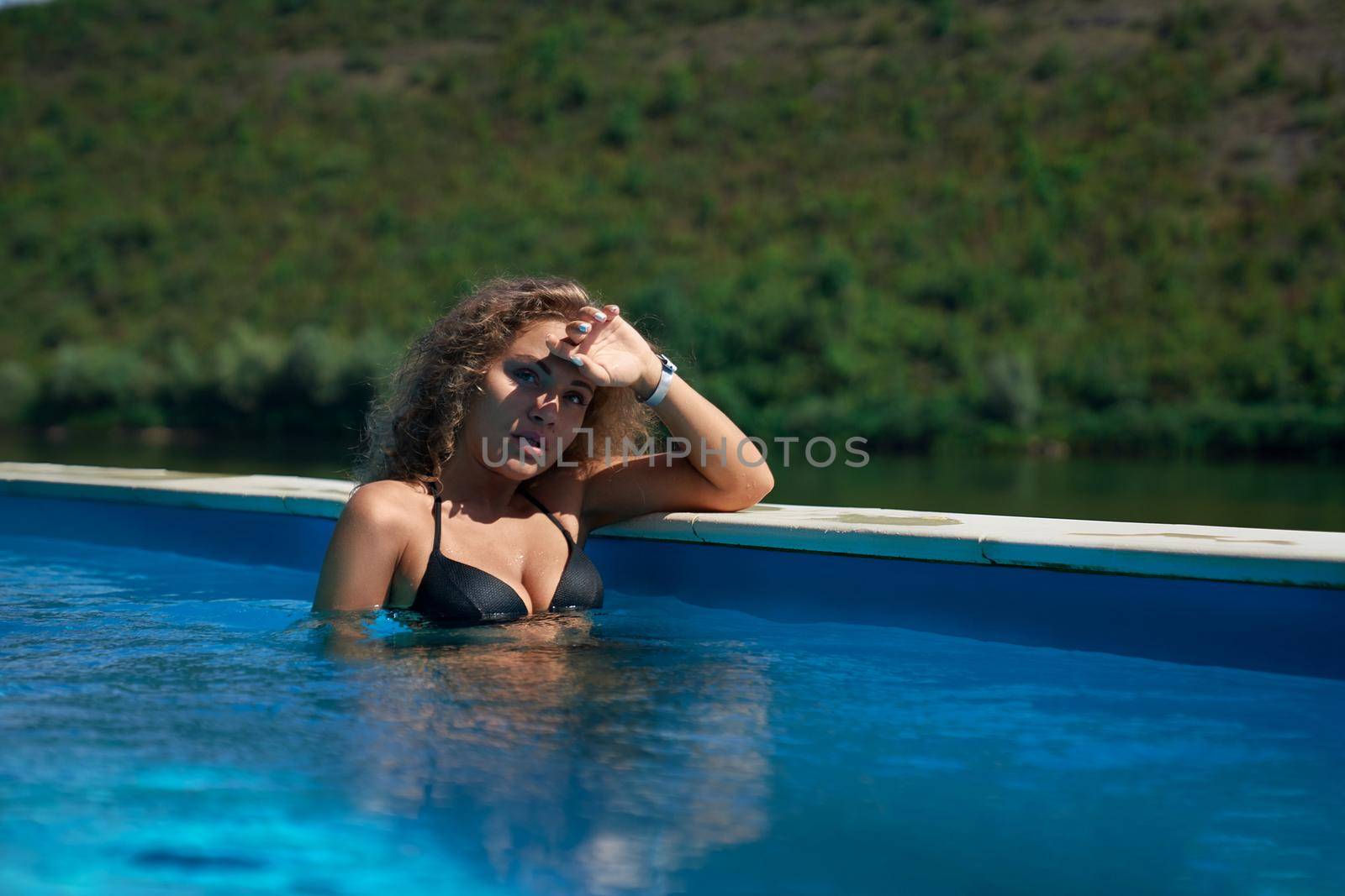 Beautiful, seductive model relaxing in swimming pool of spa resort and leaning on edge of pool. Sexy girl looking at camera and hiding her hand from sun. Concept of comfortable stay.