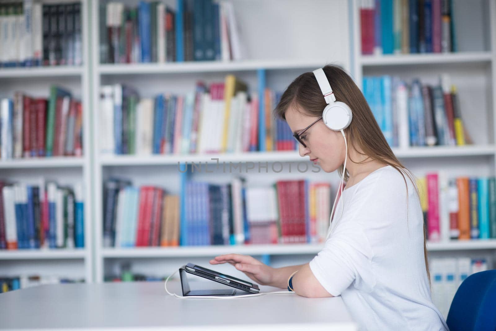 female student study in school library, using tablet and searching for information’s on internet. Listening music and lessons on white headphones