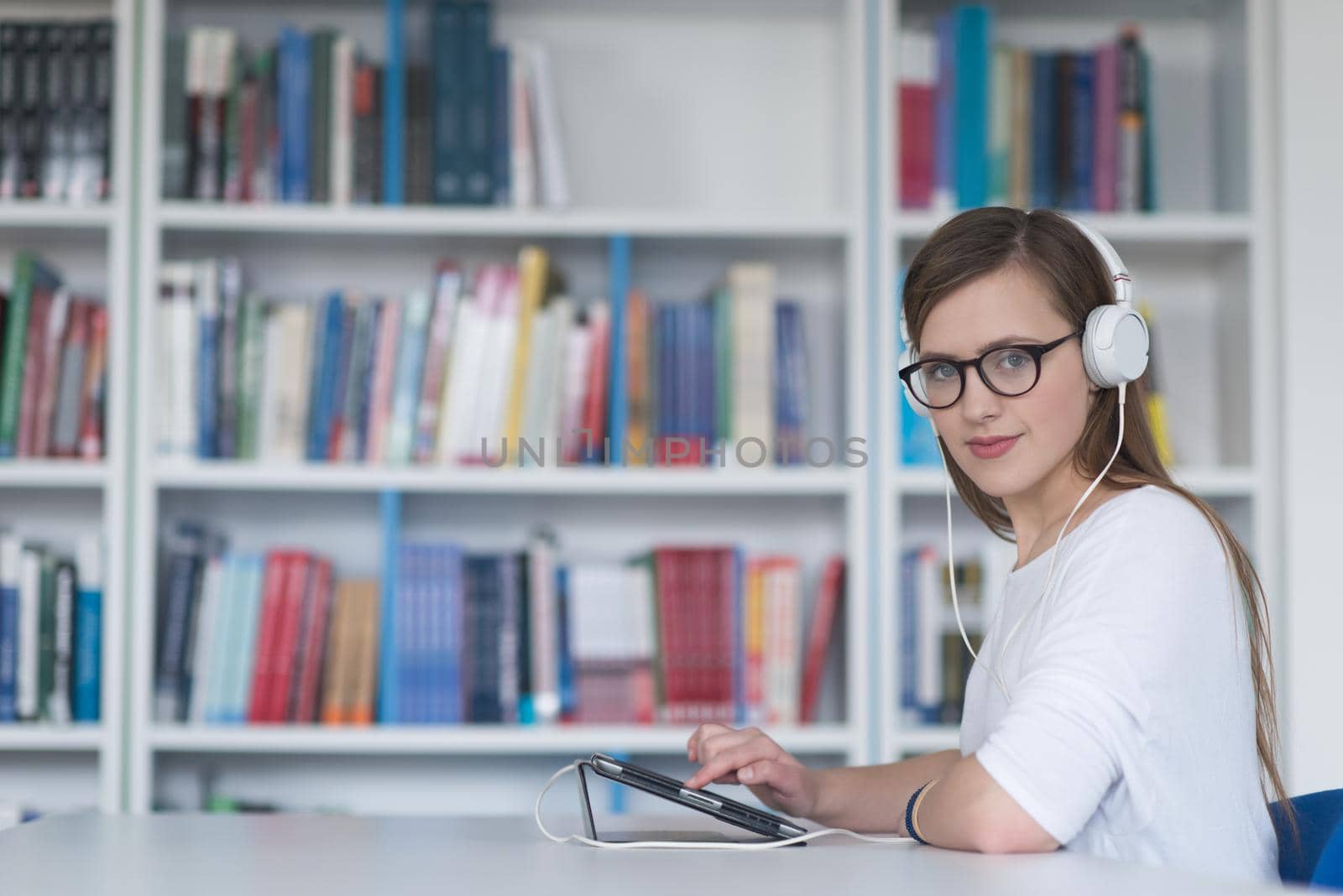 female student study in school library, using tablet and searching for information’s on internet. Listening music and lessons on white headphones