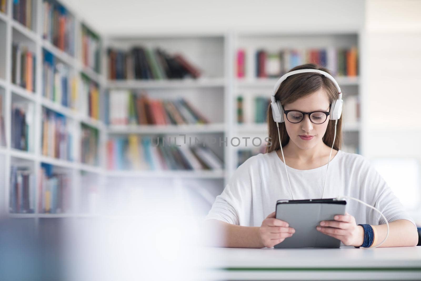 female student study in school library, using tablet and searching for information’s on internet. Listening music and lessons on white headphones
