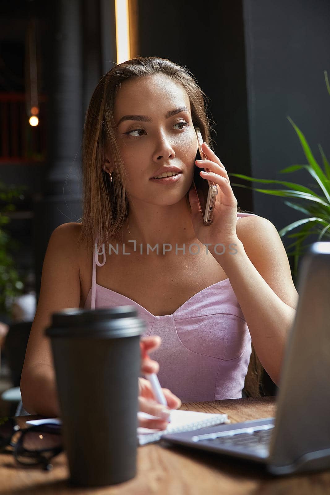 beautiful young asian woman sitting with laptop at cafe, calling by smartphone by artemzatsepilin