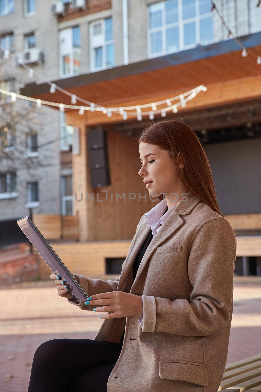 young caucasian woman in coat using tablet outdoors on sunny day, reading ebook, watching movie. attractive female street portrait at fall or spring. autumn lifestyle, modern communication technology