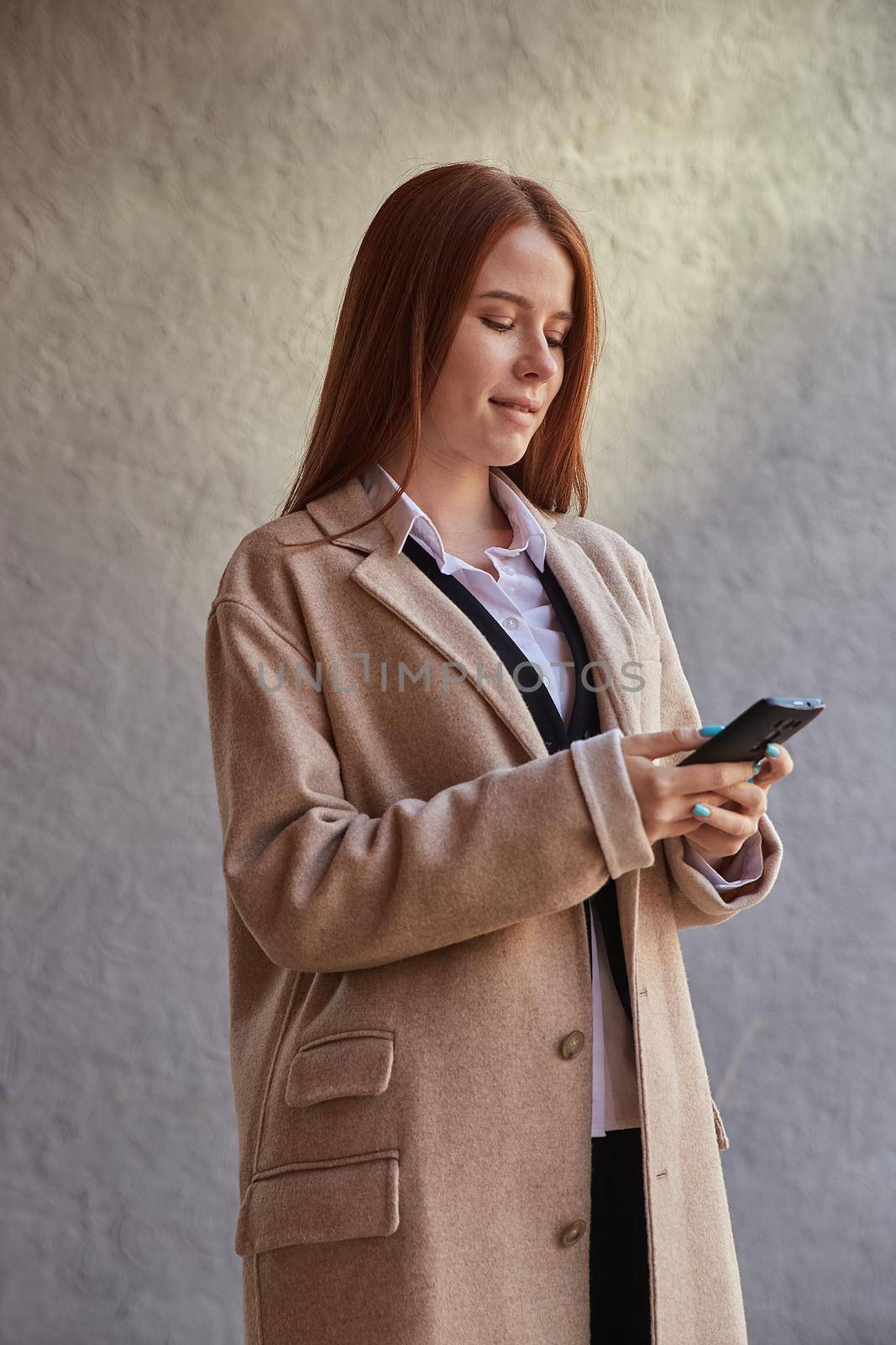 young caucasian woman in coat using smartphone outdoors on sunny day, reading news, social media. attractive female street portrait at fall or spring. autumn lifestyle, modern communication technology