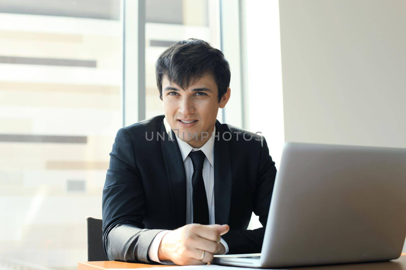 Portrait of happy businessman sitting at office desk, looking at camera, smiling.