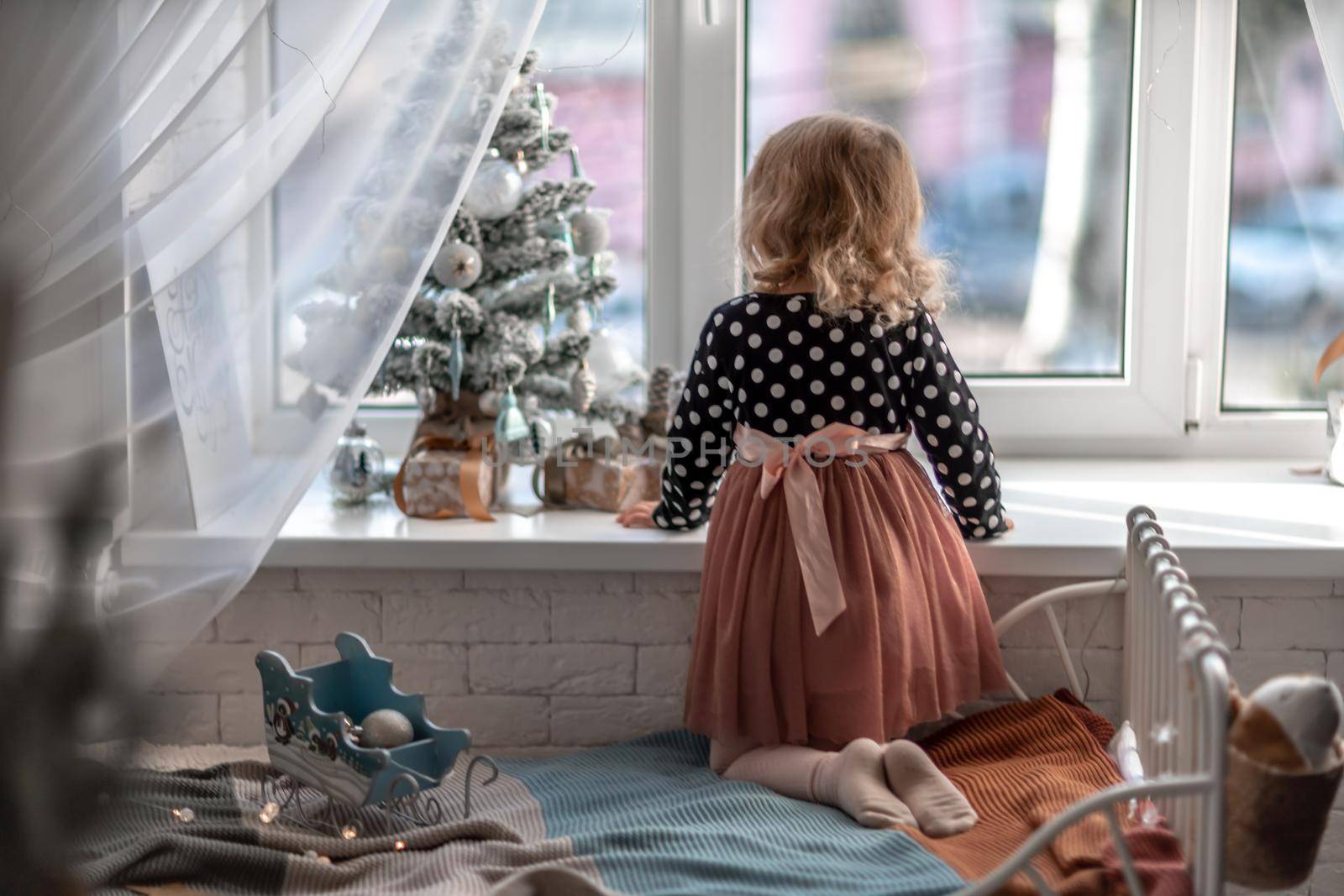 A little girl is sitting on the bed by the window and decorating a small tree with tiny Christmas toys. Happy healthy child celebrating a traditional family holiday. Adorable baby