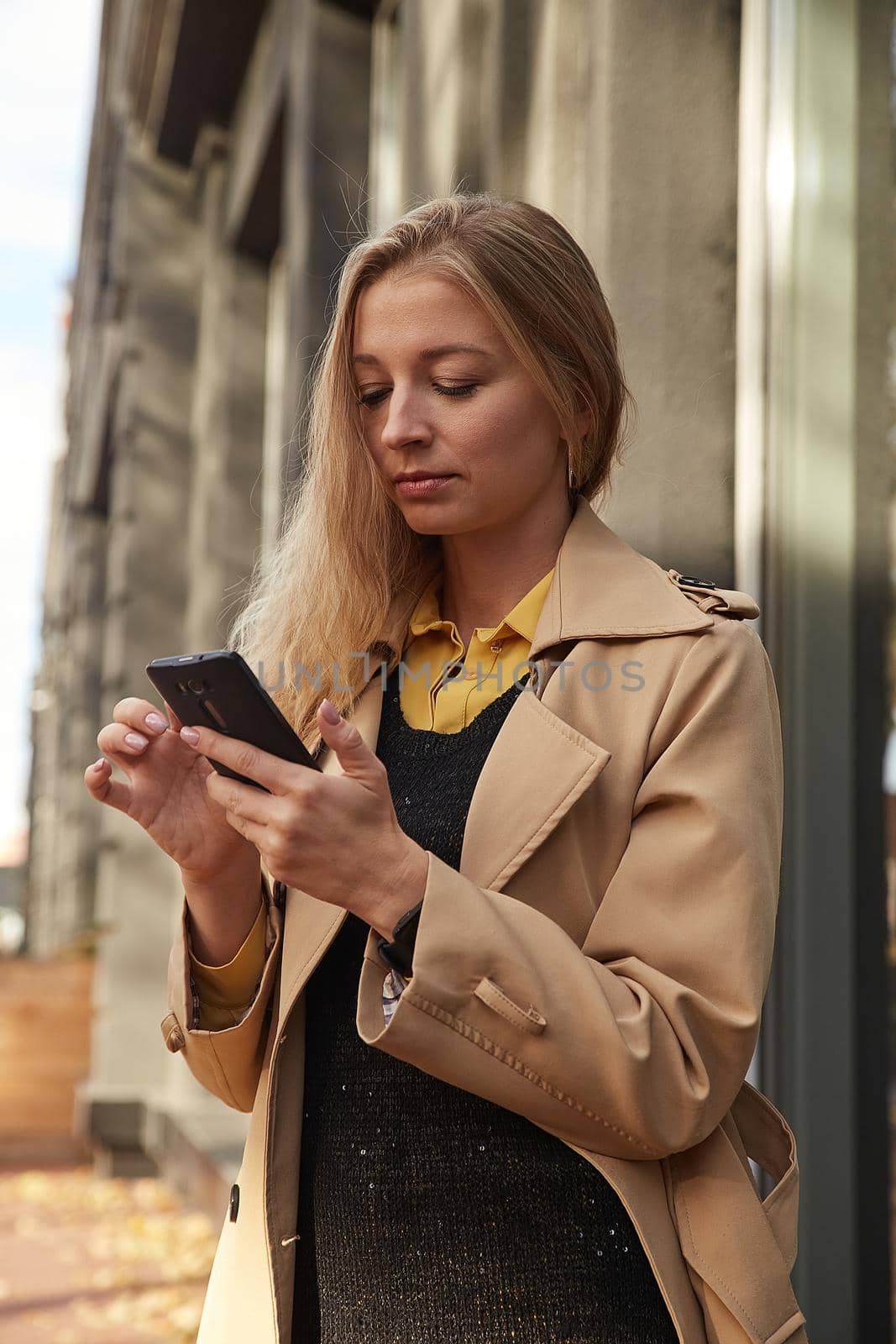 caucasian woman in beige trench coat using smartphone outdoors on sunny day, reading news, social media. attractive female street portrait at fall or spring. autumn lifestyle, modern communication