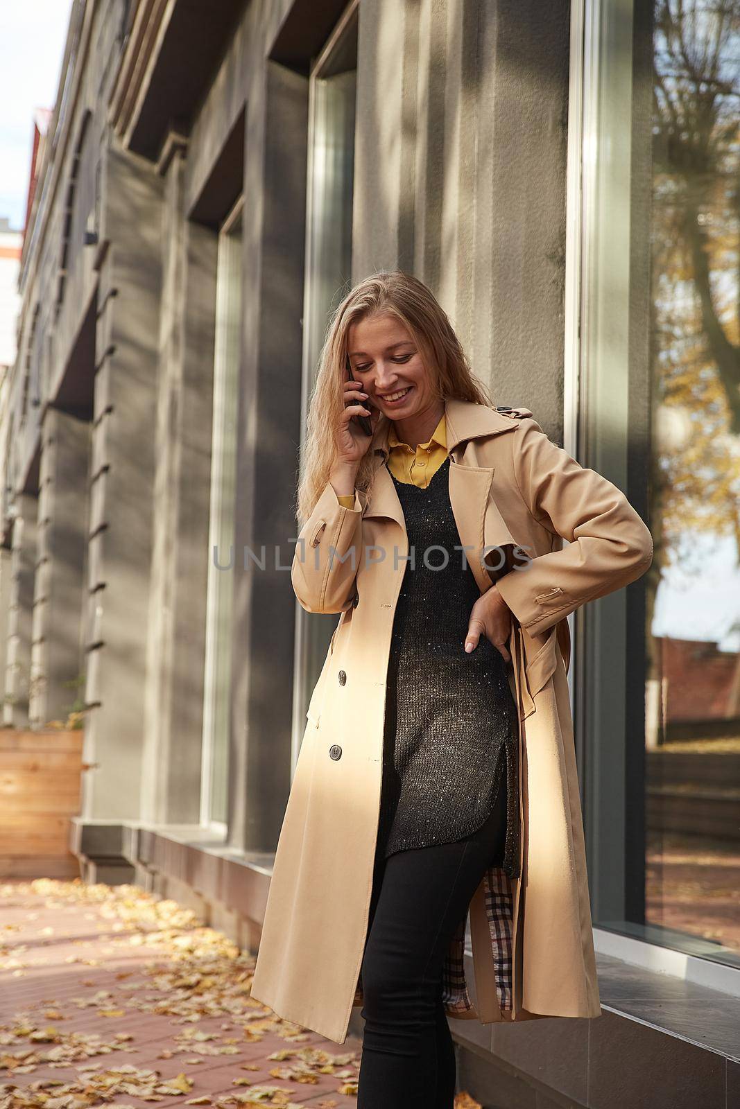 caucasian woman in beige trench coat calling by smartphone outdoors on sunny day by artemzatsepilin