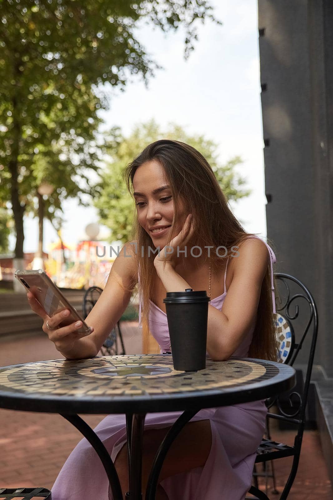 young pretty woman sitting at table with beverage, using her smartphone. lady having cup of coffee outside of cafe, surfing internet on cell phone, chatting online. modern communication technology