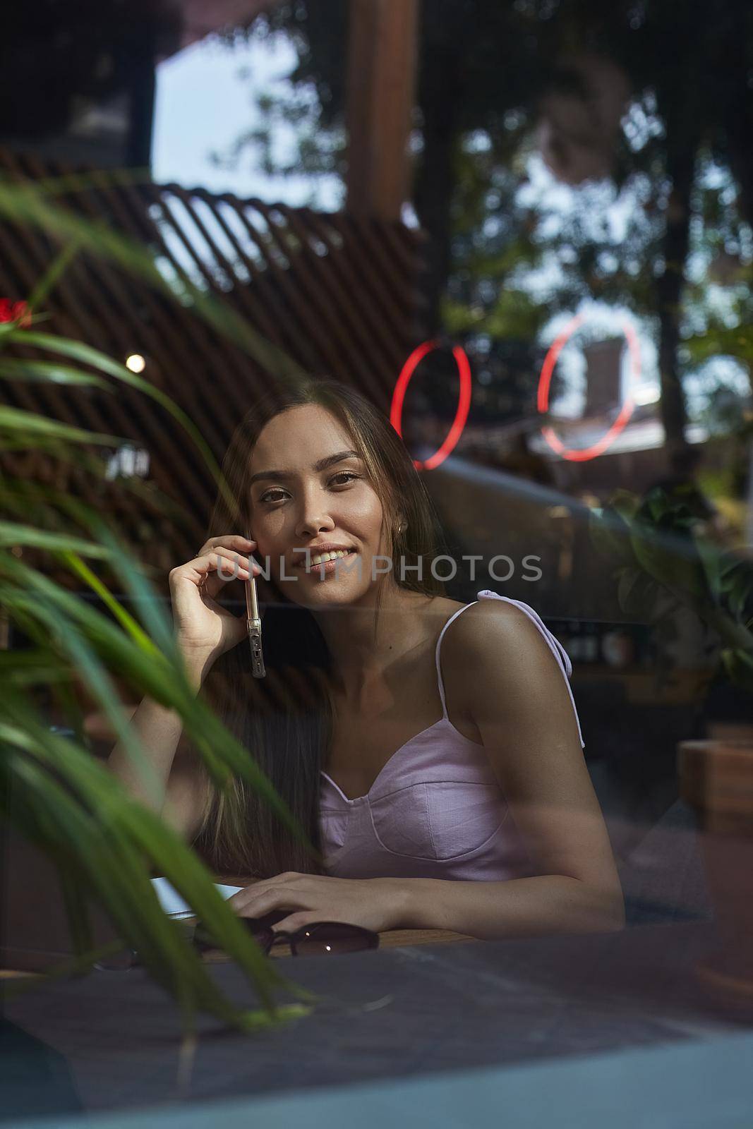 young asian pretty woman sitting at table of coffee shop, calling by smartphone. beautiful smiling lady using cell phone shot through window glass outside of cafe. modern communication technology