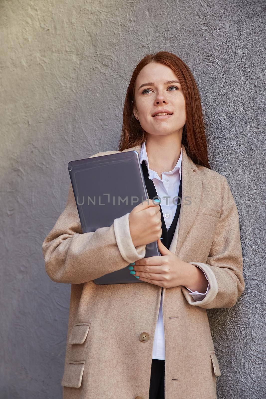 young caucasian stylish pretty girl in beige coat holding tablet outdoors by artemzatsepilin