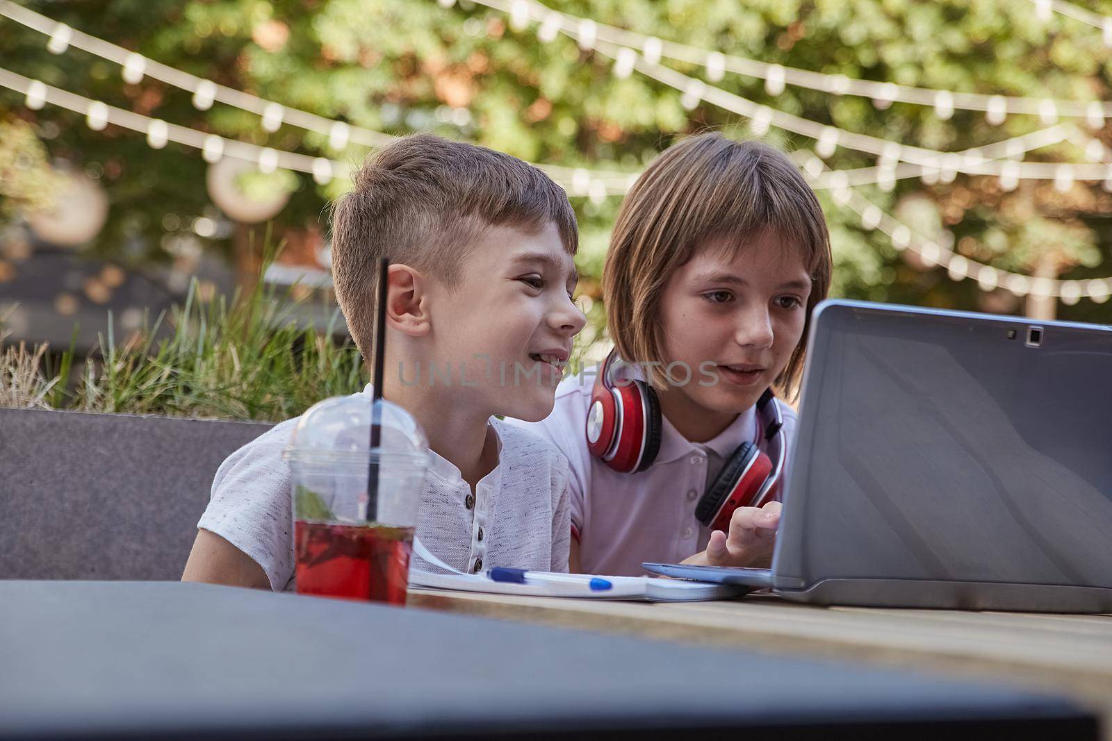 boy and girl sit outdoors, watch at screen of laptop. little caucasian schoolchildren sitting at table at veranda of cafe, studying with computer, watching movie. distant learning, online education