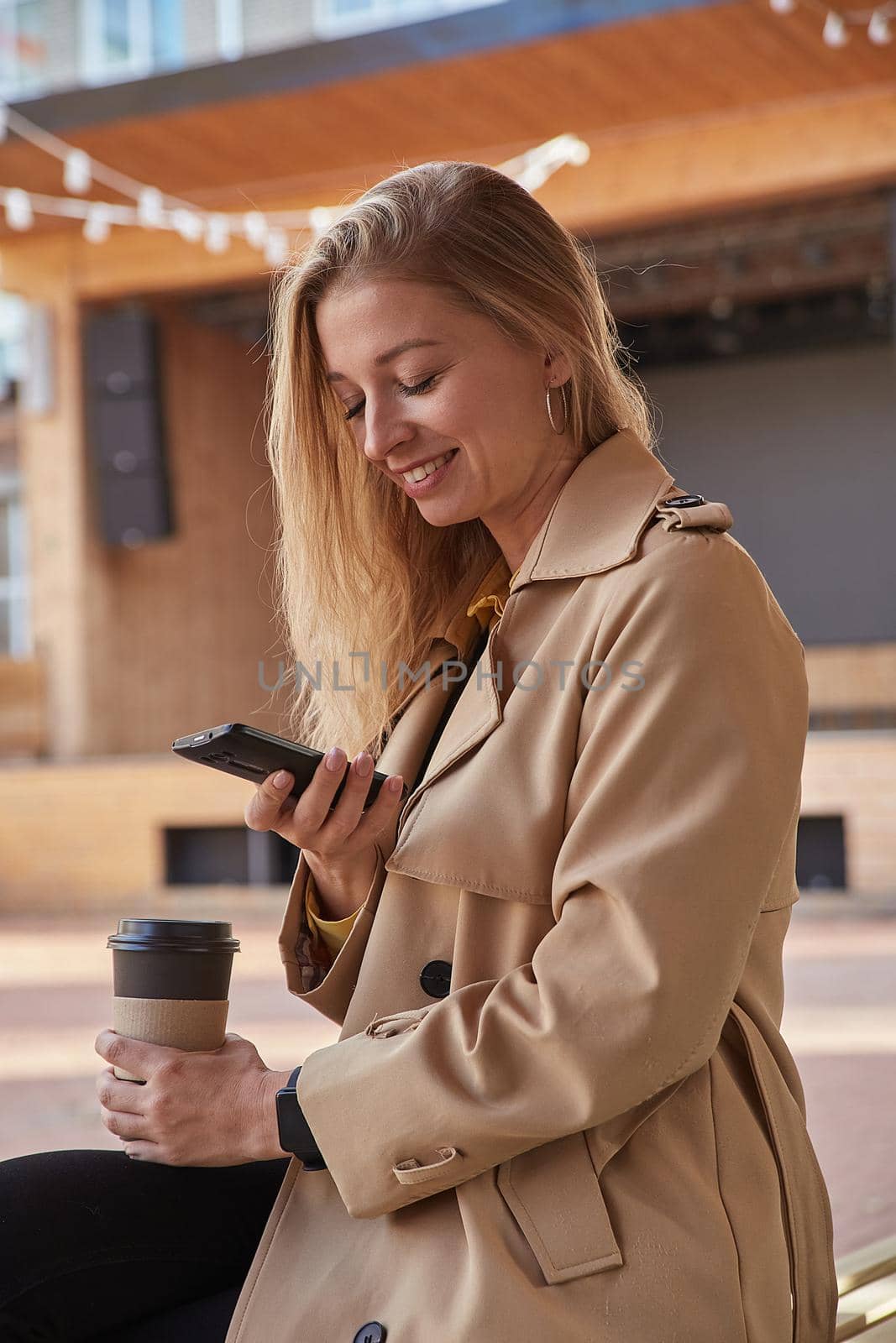 caucasian woman in beige trench coat using smartphone outdoors on sunny day, reading news, social media. attractive female street portrait at fall or spring. autumn lifestyle, modern communication