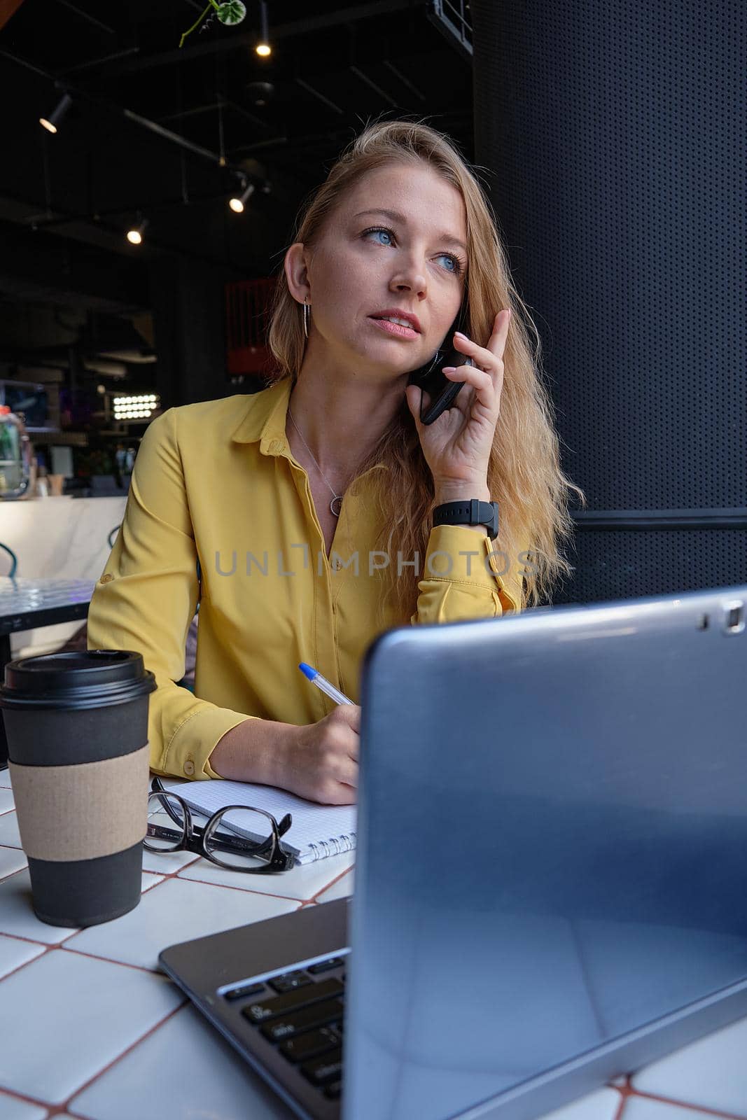 young cauasian pretty woman sitting with laptop at cafe, calling by smartphone by artemzatsepilin