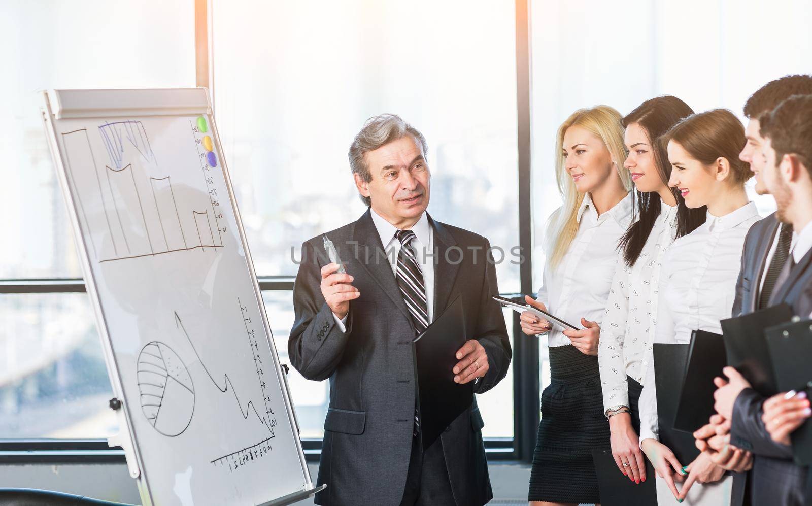 Head of business team explains to his colleagues the methods of sales growth, using a stand with charts and graphs in a modern office