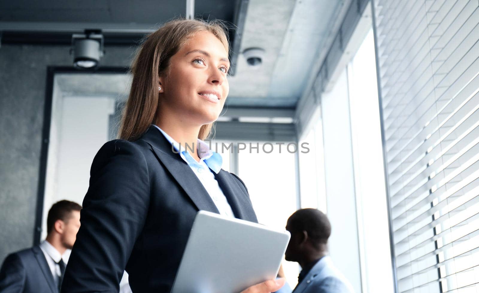 Business woman standing in foreground with a tablet in her hands, her co-workers discussing business matters in the background by tsyhun