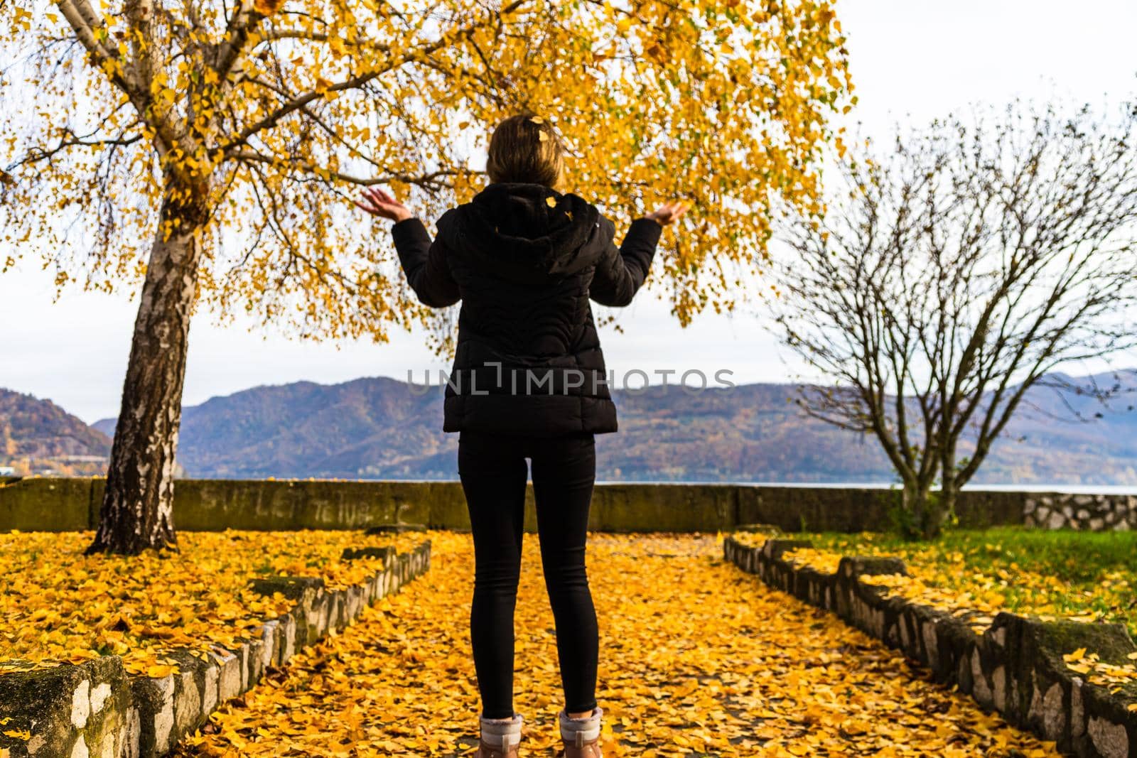 Autumn leaves fallen on alone woman walking on the autumn alley. Autumn landscape, orange foliage in a park in Orsova, Romania, 2020