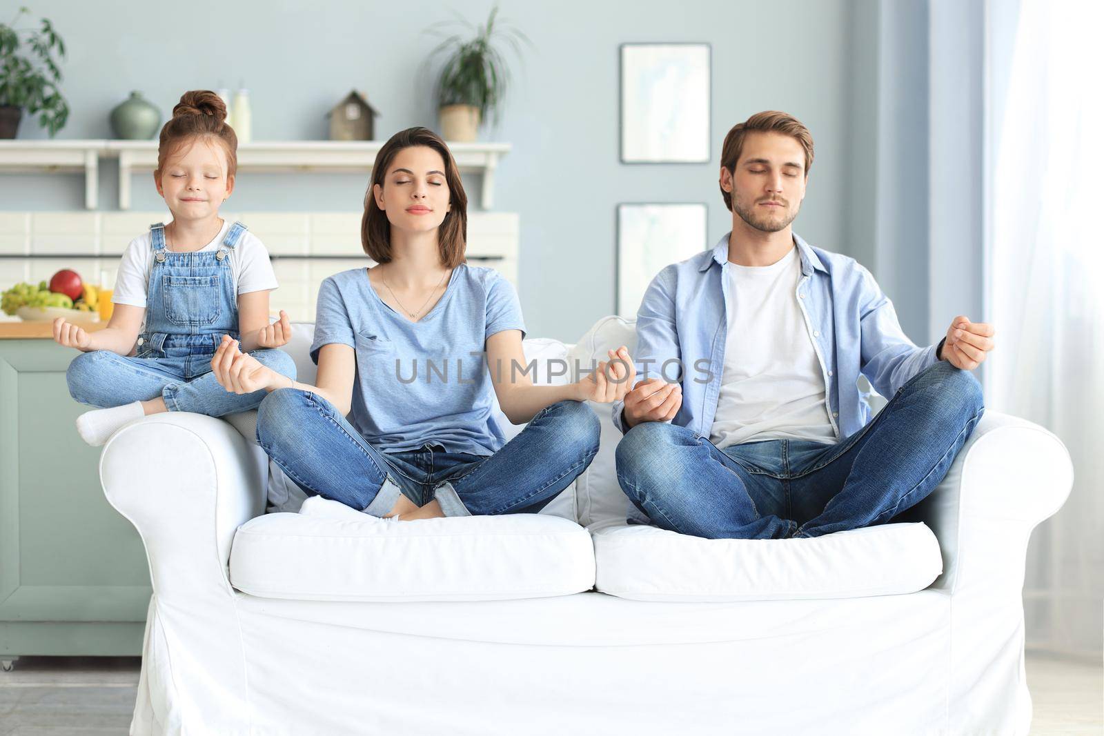 Calm young family with little daughter sit on couch practice yoga together, happy parents with small preschooler girl child rest on sofa meditate relieve negative emotions on weekend at home by tsyhun