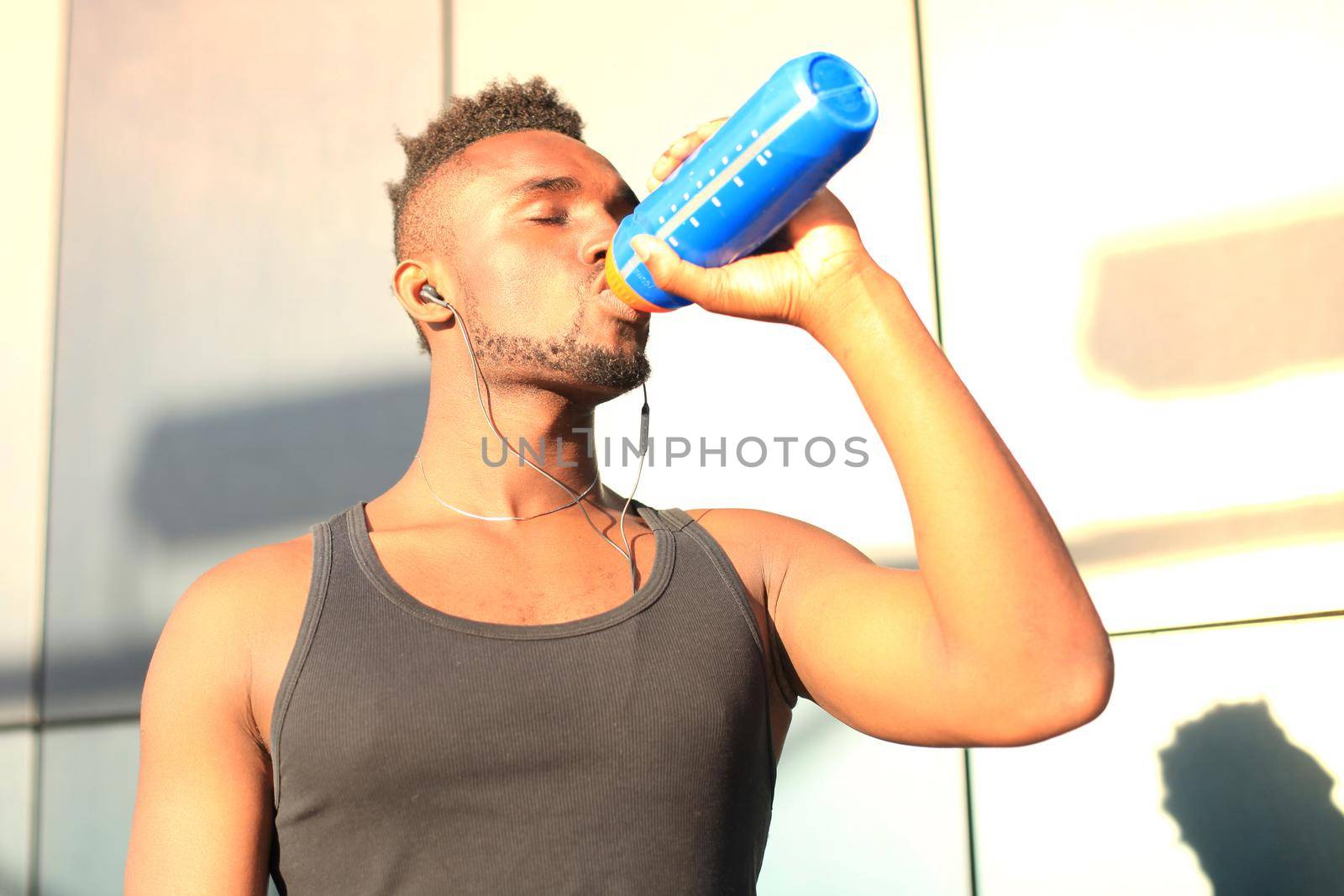 African man in sports clothing drinking water while standing outside, at sunset or sunrise. Runner. by tsyhun