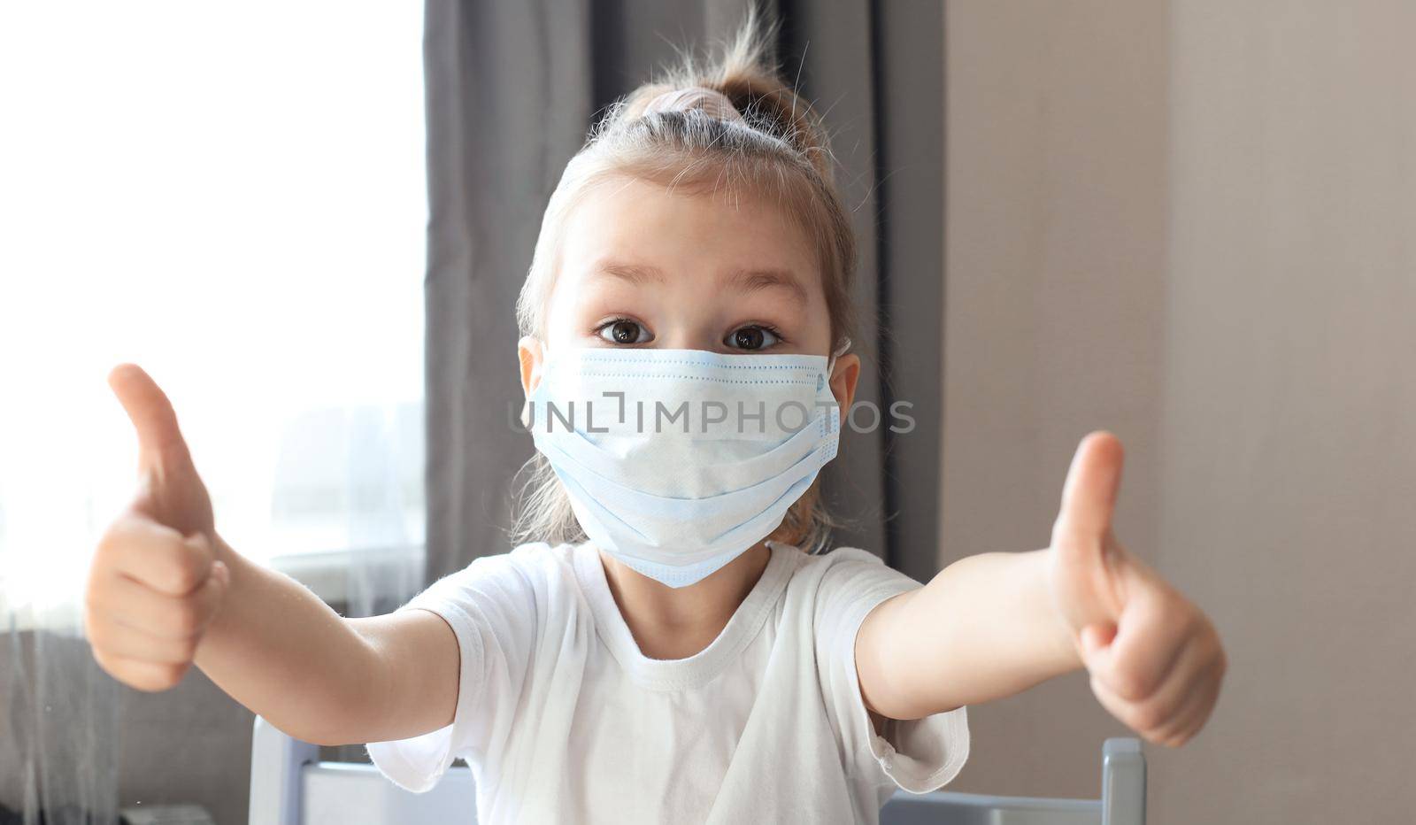 Little girl in a medical mask showing thumb's up gesture. Protection against coronavirus