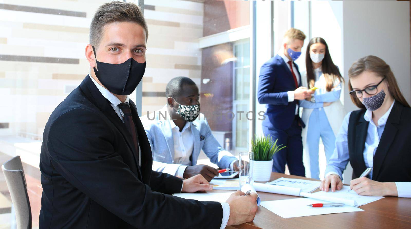 Businessman in protective mask sitting in the office with his colleagues on a background.