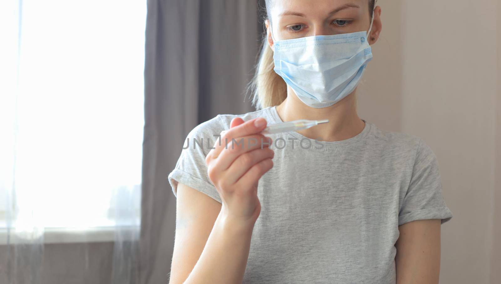 Young woman in the medical mask looks at the thermometer with high temperature. by tsyhun