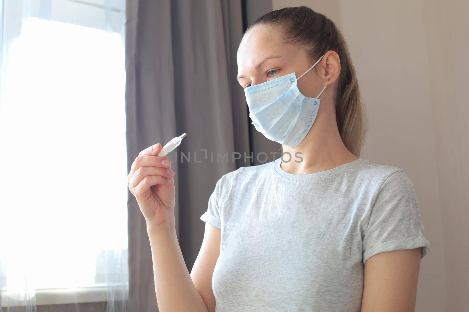 Young woman in the medical mask looks at the thermometer with high temperature. by tsyhun