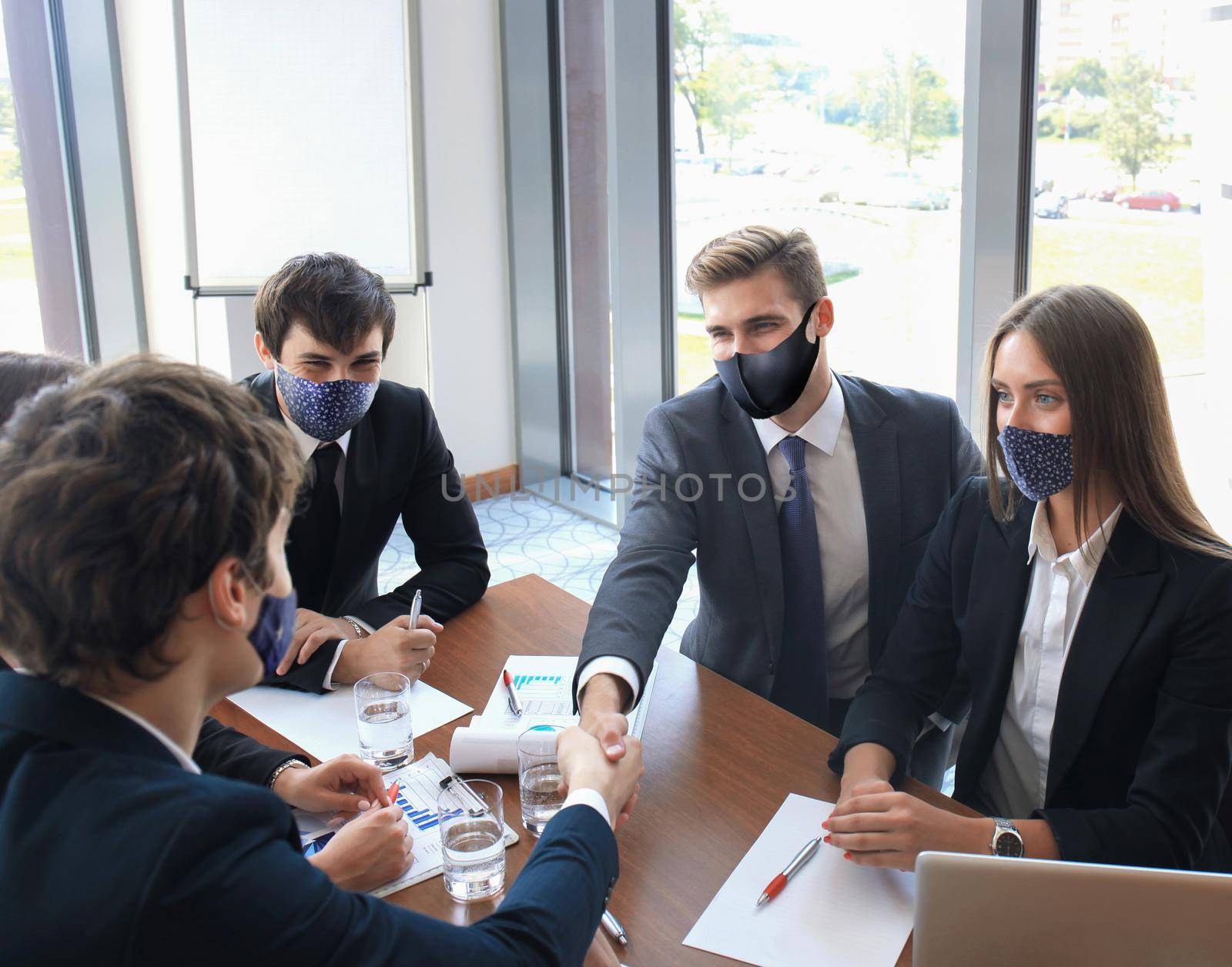 Businessman in preventive mask shaking hands to seal a deal with his partner and colleagues who also wear preventive masks in office. by tsyhun