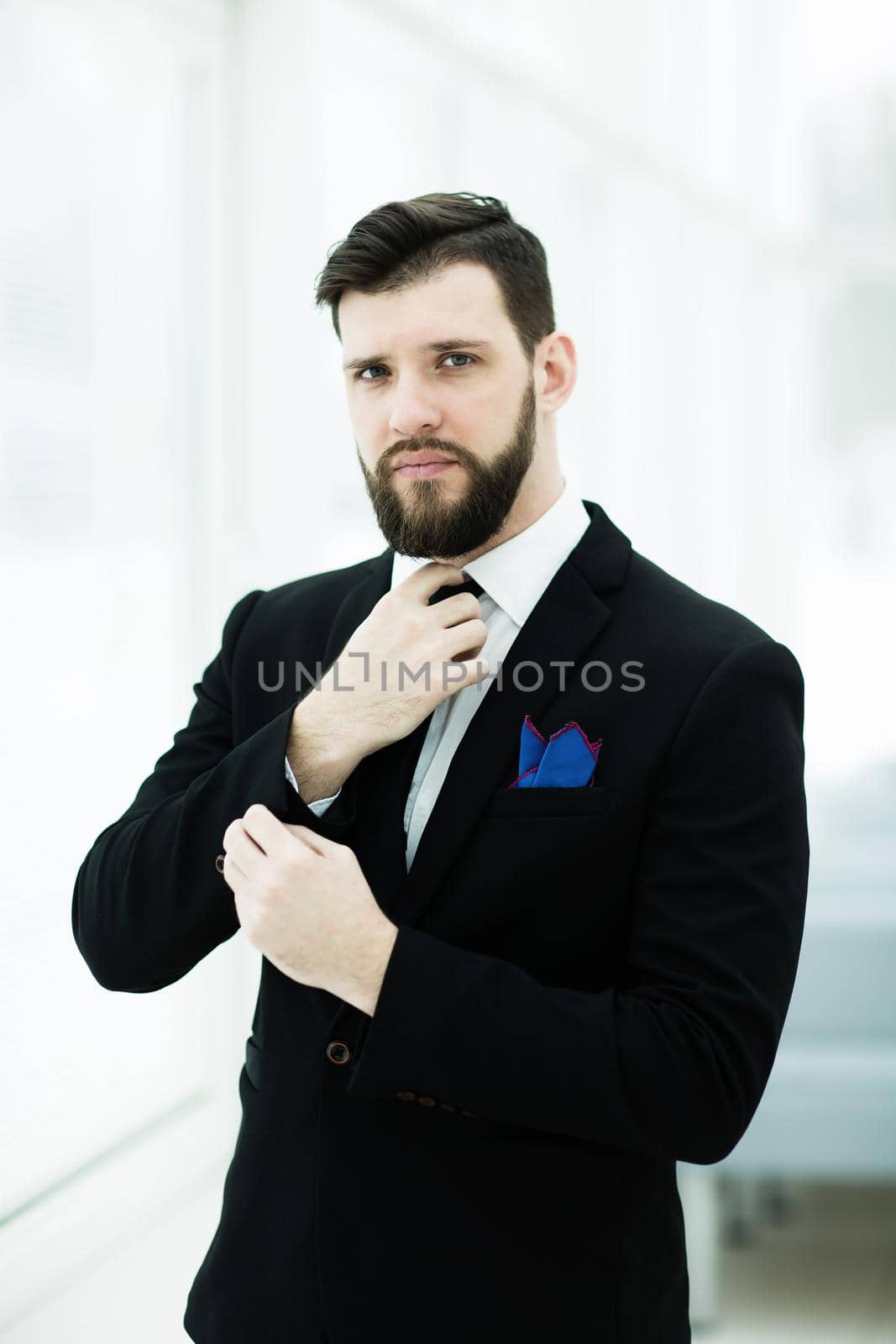 portrait of a successful businessman adjusting his necktie while standing near a window in bright office.the photo has a empty space for your text