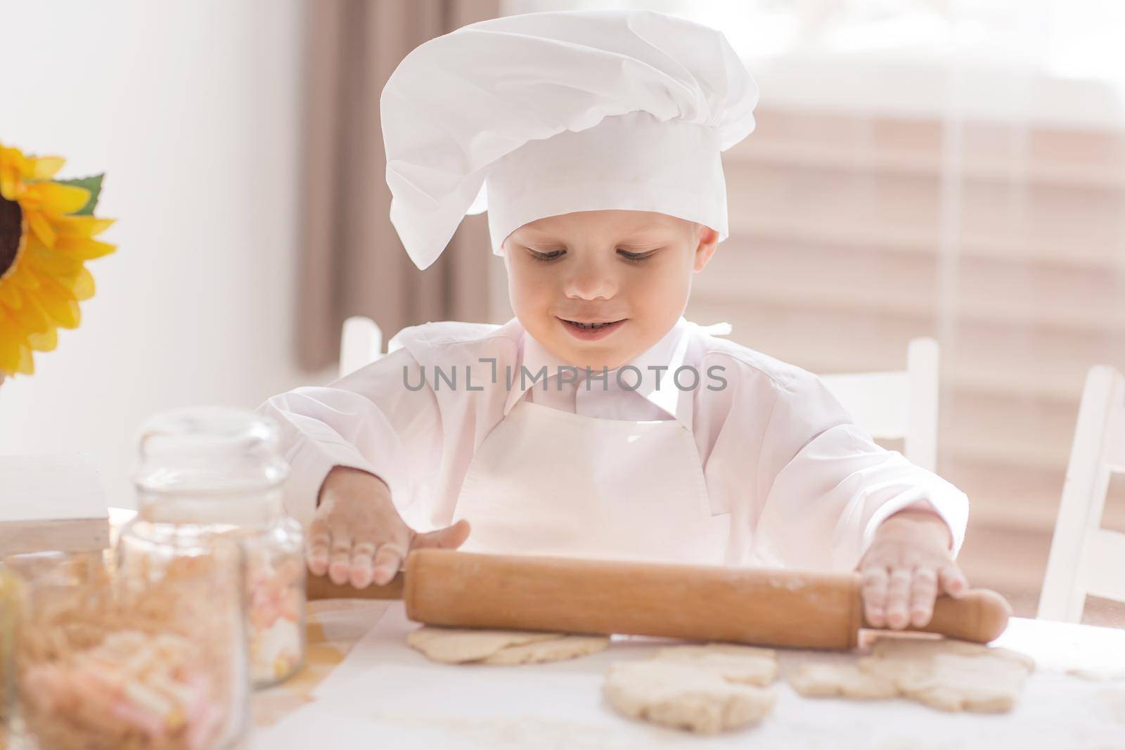 A small child in the form of a cook rolls out the dough on the kitchen table