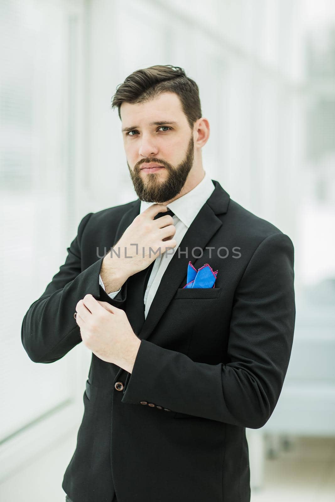 portrait of a successful businessman adjusting his necktie while standing near a window in bright office.the photo has a empty space for your text
