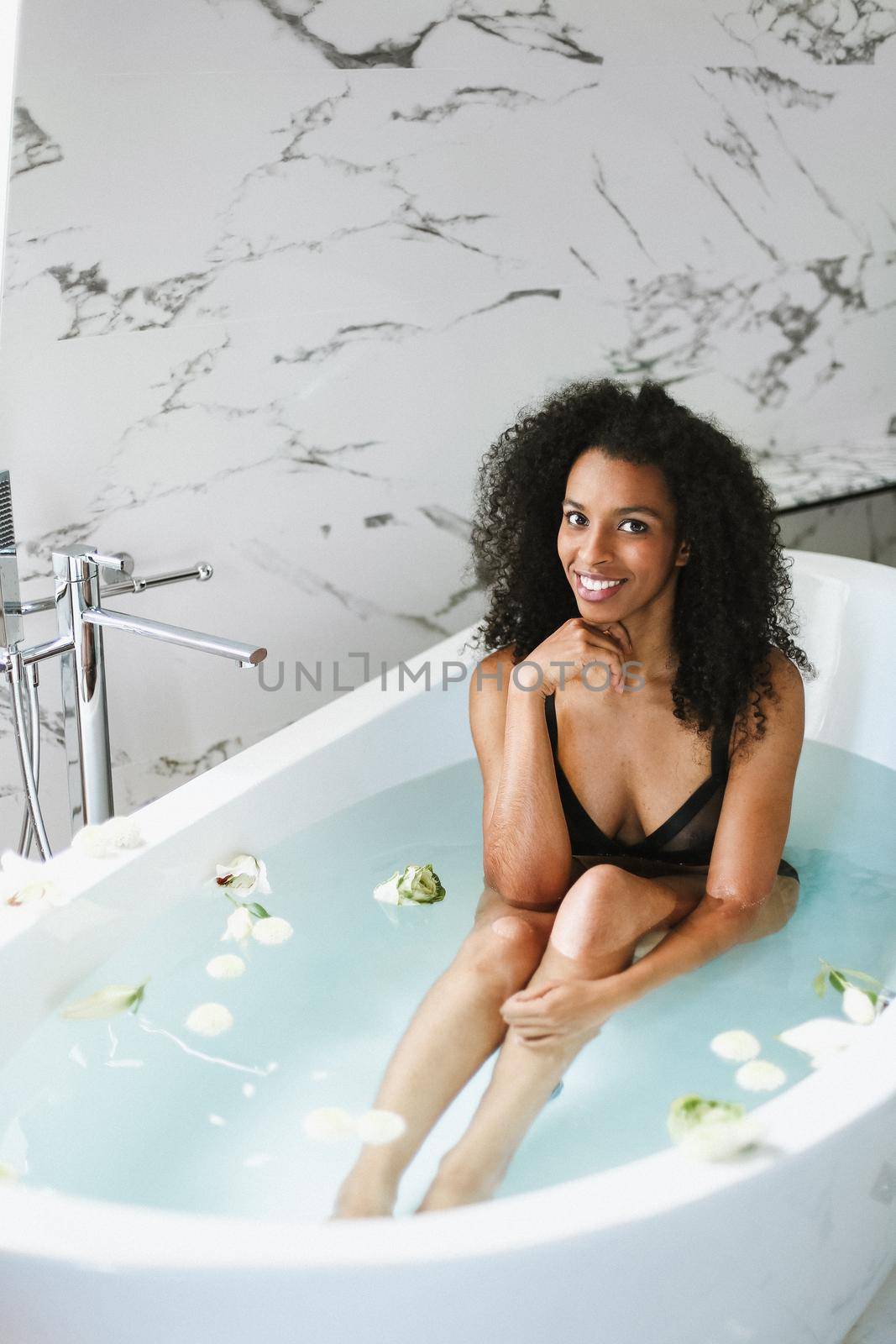 Young afro american girl sitting in bathroom with marble tile, wearing black swimsuit. Concept of personal care and spa.