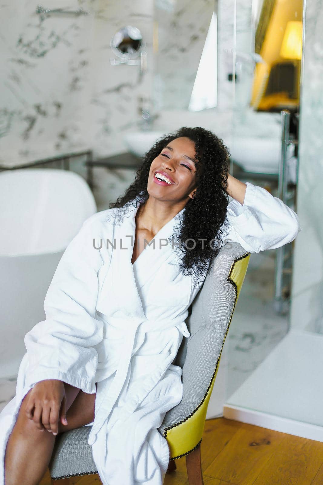 Brown skin relaxed girl with curly hair sitting in bathroom and wearing white bathrobe. by sisterspro