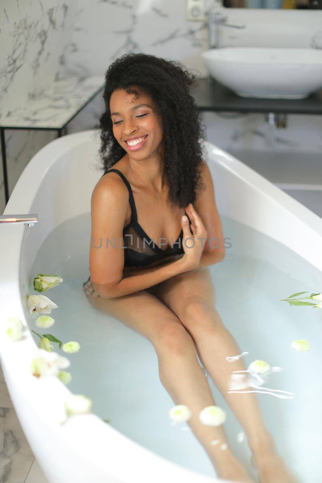 Afro american smiling girl lying in bath with flowers and wearing black bra and panties. Concept of relaxing and bathroom photo session in underwear.