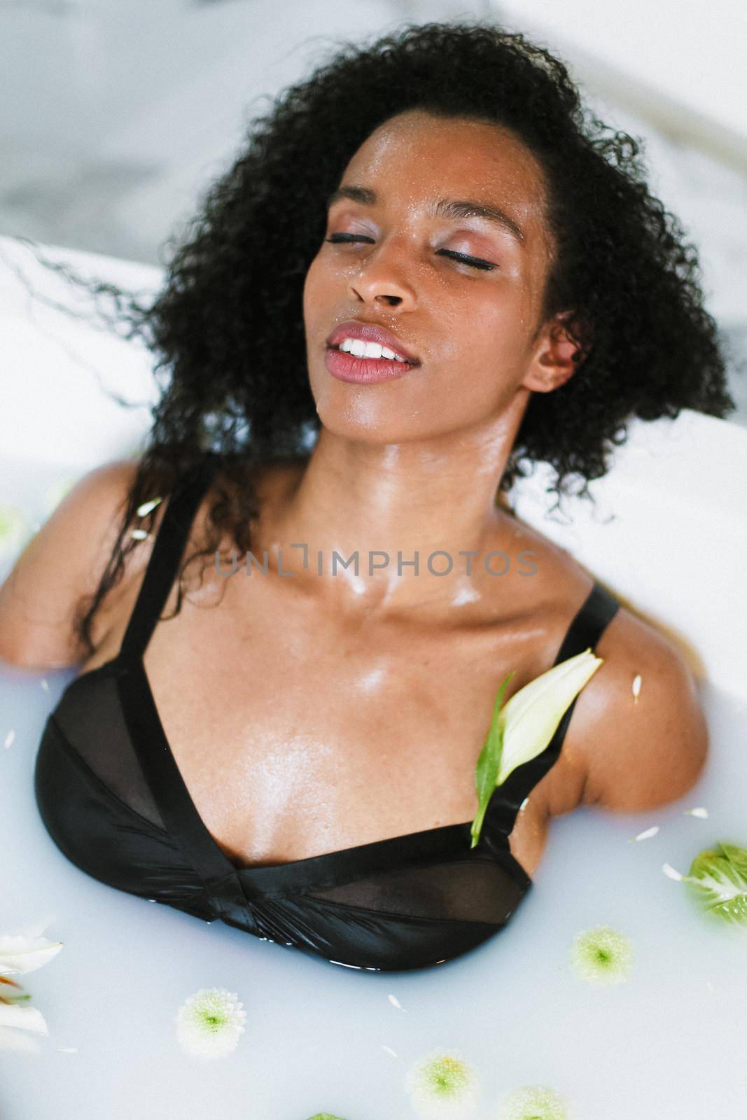 Young brown skin female person relaxing in bath with flower on shoulder, wearing black swimsuit. by sisterspro