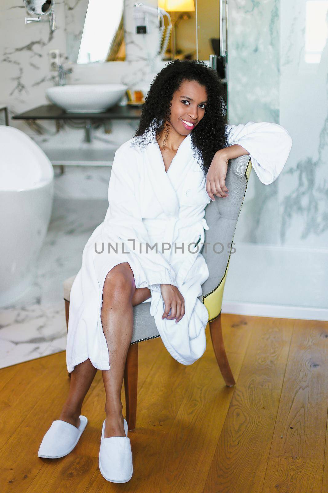 Brown skin girl with curly hair sitting in bathroom and wearing white bathrobe. by sisterspro