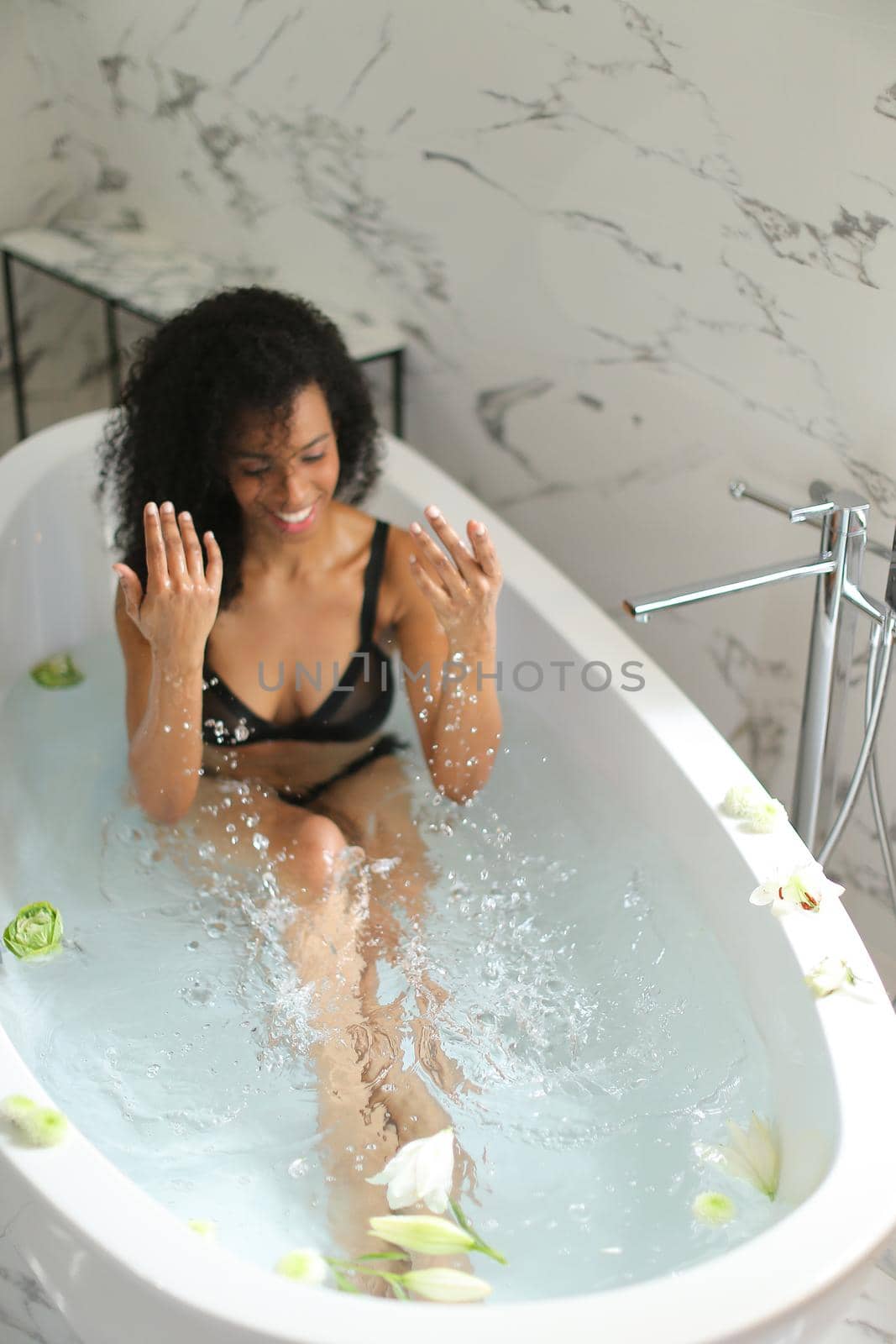 Young nice afro american woman playing with splashing water in bathroom and wearing black swimsuit. oncept of taking bath with flowers.