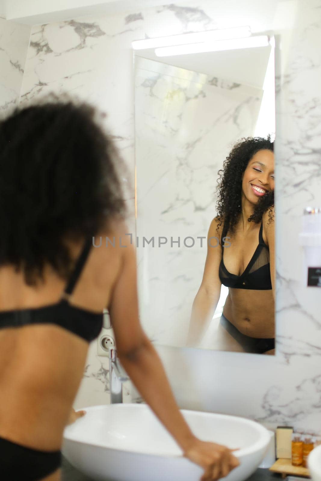 Back view afro american curly haired happy girl looking at mirror reflection in bathroom and wearing panties with bra. Concept of morning hygiene and underwear photo session.