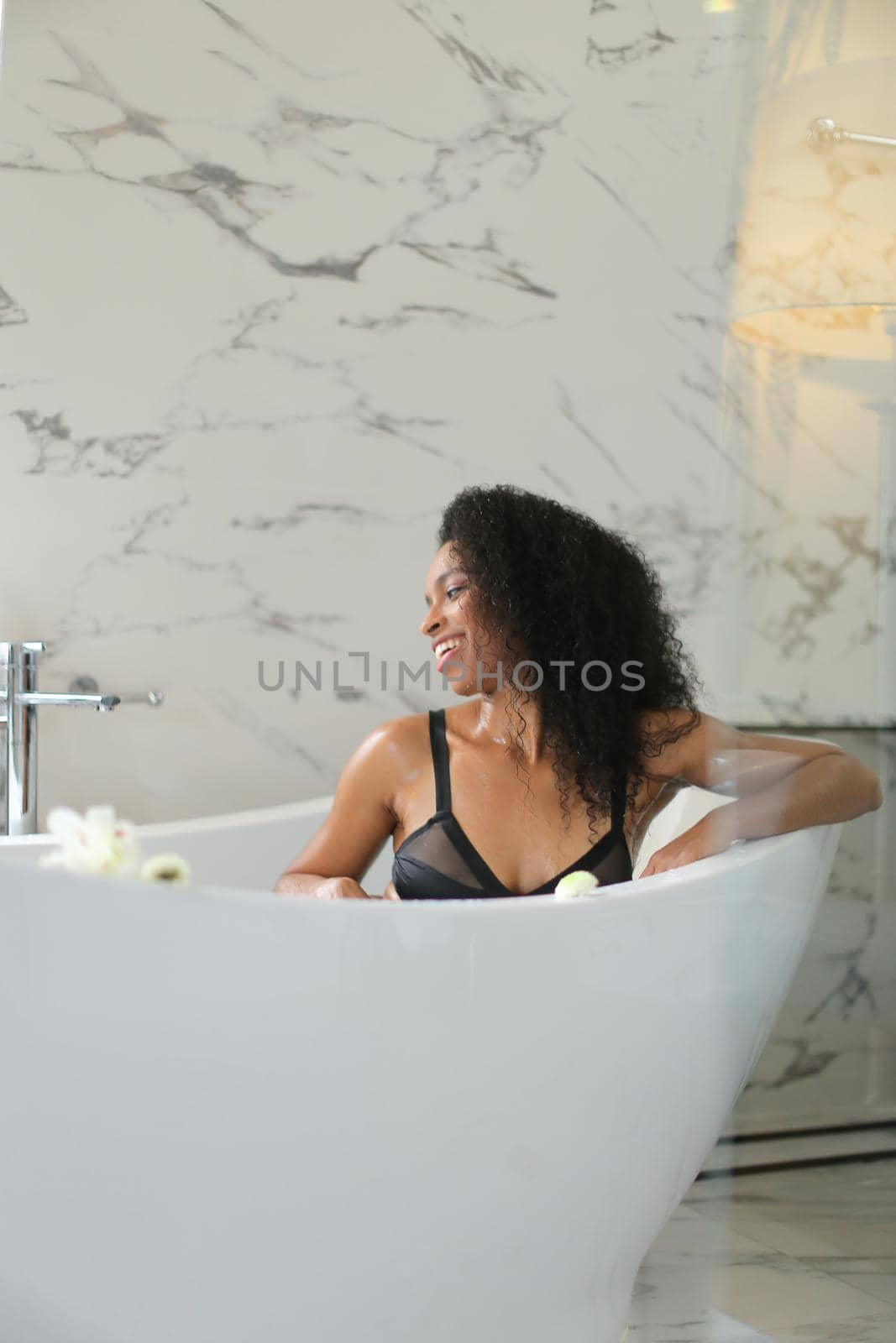 Afro american woman sittling in bath and wearing black swimsuit, marble wall in background. Cocept of bathroom photo shoot and morning hygiene.