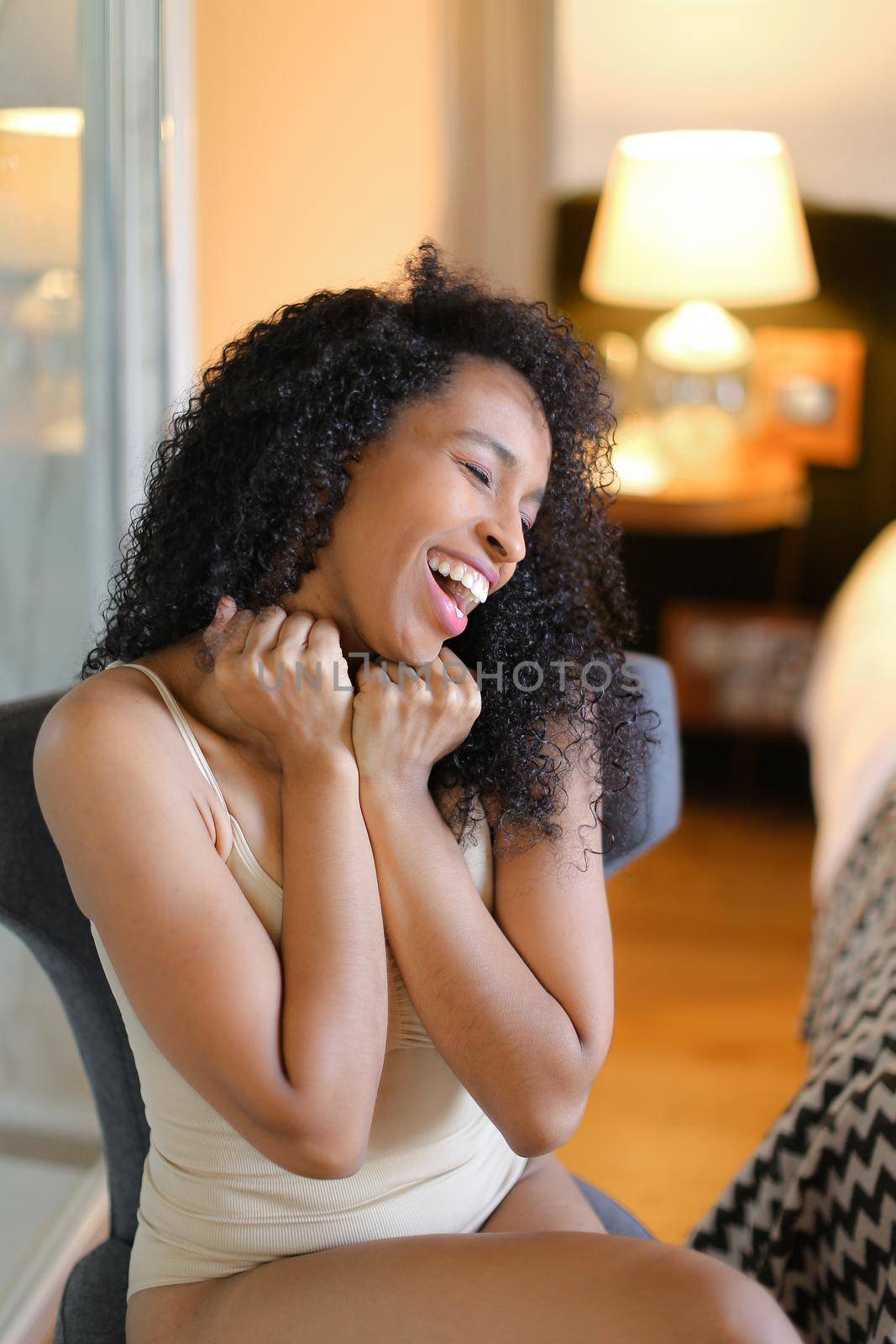 Young gladden afro american woman sitting in room and wearing beige swimsuit. Concept of hotel photo session in lingerie.