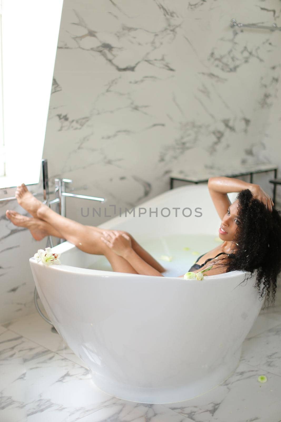 Young beautiful afro american woman taking bath and wearing black swimsuit. Concept of milk bathroom photo shoot and morning hygiene.