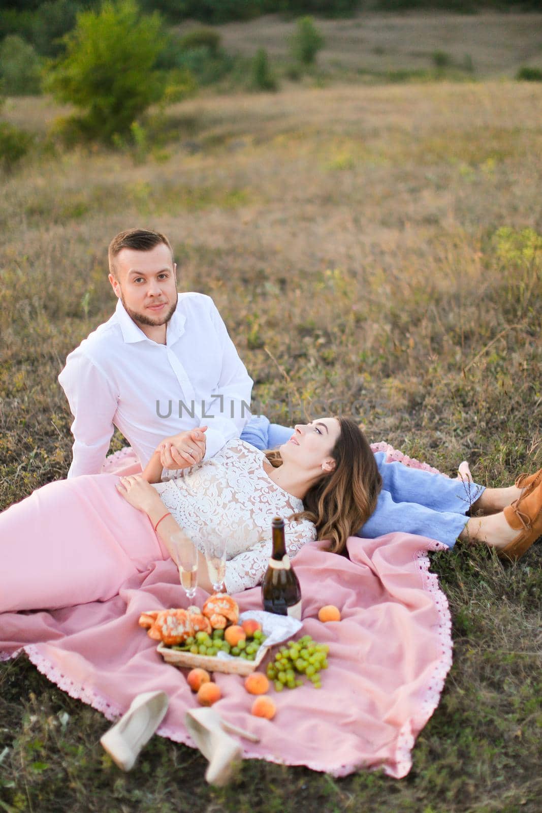 Young girl lying on man knees and pink plaid in steppe near fruits and bottle of red wine. oncept of picnic and relationship, romantic love.