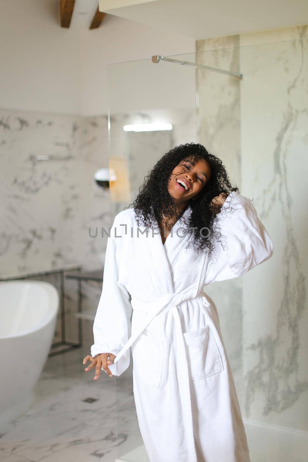 Young black girl wearing white bathrobe and standing in hotel bathroom with marble walls. Concept of relax and morning hygiene.