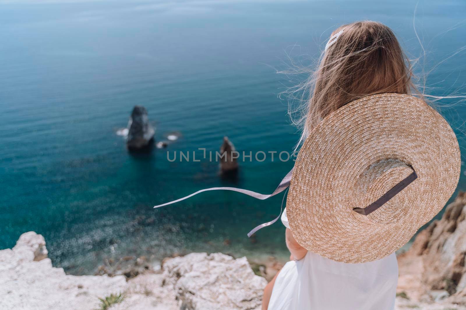 A young woman in a white dress and hat stands with her back on a rock and looks at the sea. Rocks are visible in the sea. The concept of romance, travel, adventure. by Matiunina