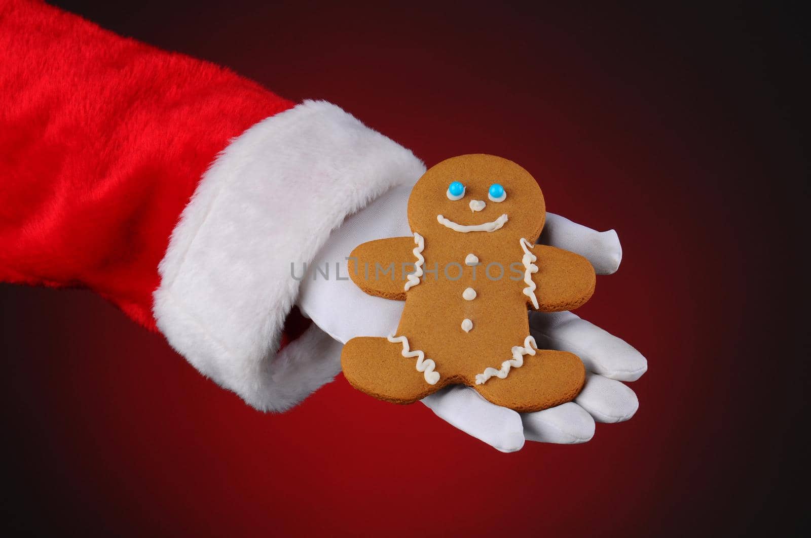 Santa Claus holding a Gingerbread man cookie in the palm of his hand. Horizontal format over a light to dark red background, showing only hand and arm.