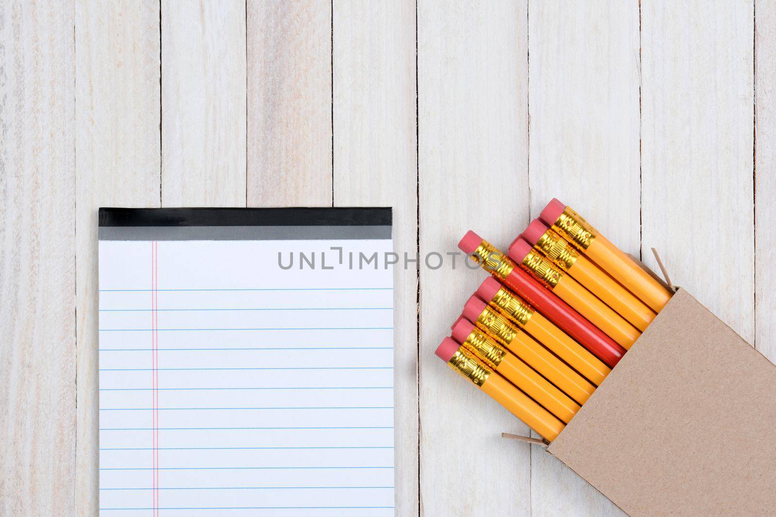 A box of yellow pencils with one red pencil partially pulled out. The plain brown box is in the lower right corner of the frame at an angle, and a blank note pad is next to the box.